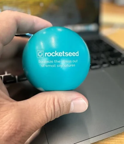 A hand holding a blue Rocketseed stress ball that says "squeeze the stress out of email signature management" with a laptop background