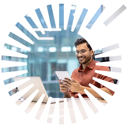 A smiling man with glasses sitting at his desk behind his laptop working on his tablet