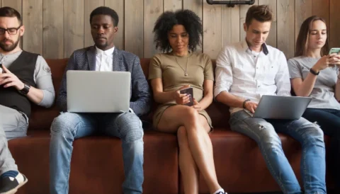 Five individuals sitting in a row on their laptops and cellphones in front of a wooden wall